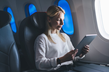 Young caucasian female passenger enjoy comfortable flight in wide seat near large controlled physical window shade with modern electrochromic technology. Tourist woman use wireless connection on board