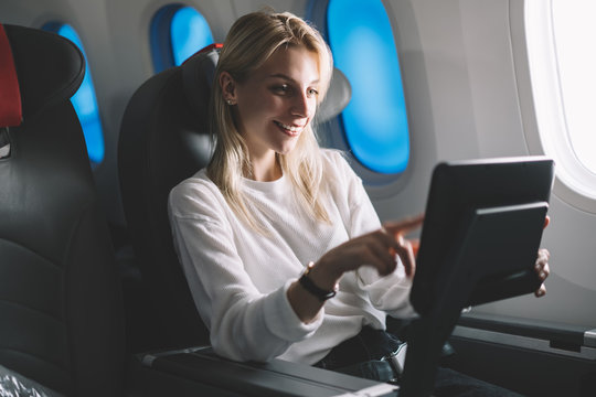 Smiling attractive female passenger searching movie to watch on board tv device during comfortable flight. Caucasian young woman traveler enjoying touch screen entertainment monitor system on airplane