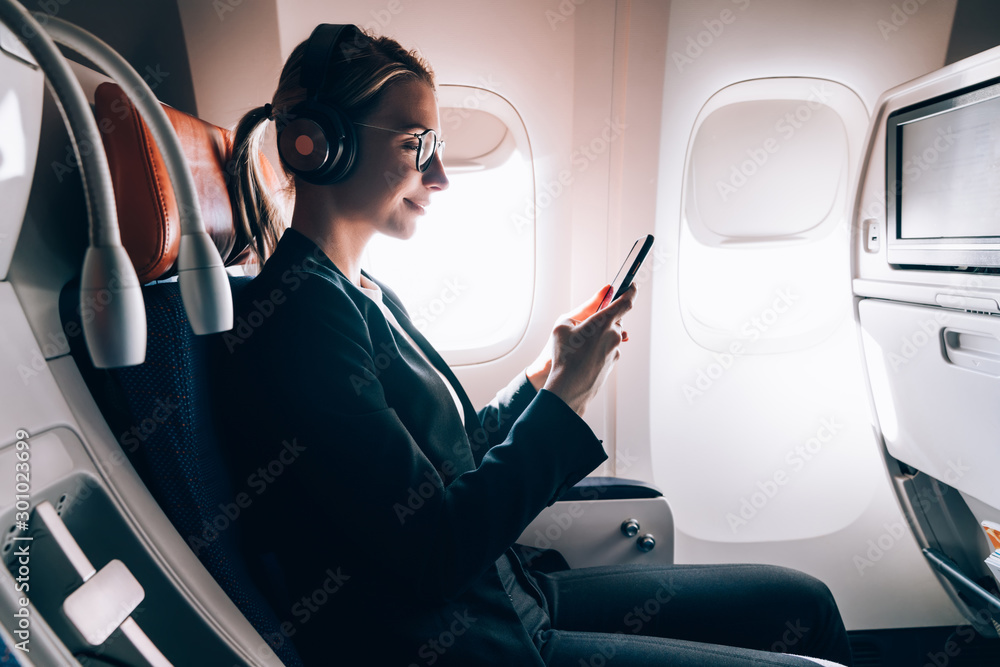 Poster smiling female passenger in bluetooth headphones for noise cancellation chatting online via mobile p