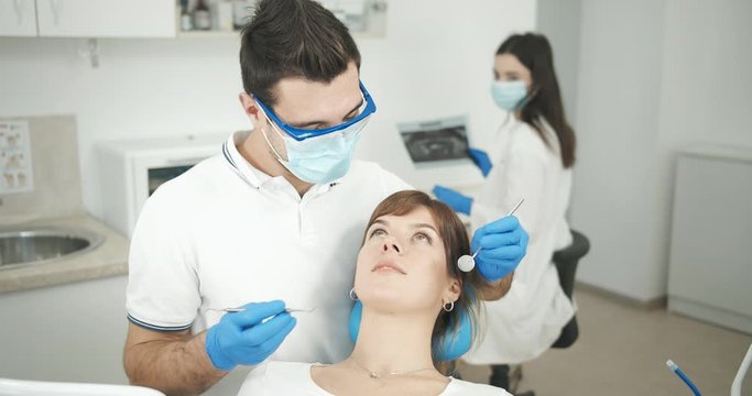 Male stomatologist in protective mask and eyeglasses checking female's teeth, nurse holding x-ray on background