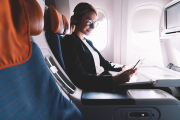 Happy smiling woman passenger in headphones for noise cancellation enjoying intercontinental flight in first class with high speed wifi connection on board using touch pad and browsing internet