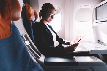 Positive caucasian woman in eyewear for vision correction reading text publications in social network during intercontinental flight connected to wifi on board, happy female browsing websites - obrazy, fototapety, plakaty