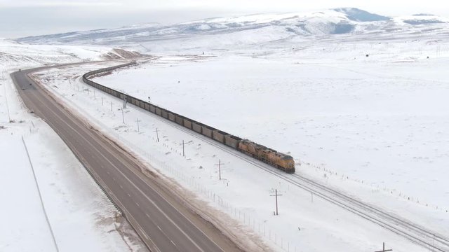 DRONE: Locomotive transports a long line of railcars full of coal across the idyllic wintry United States. Freight train moves along a highway while transporting cargo from coal mine to storage depot.