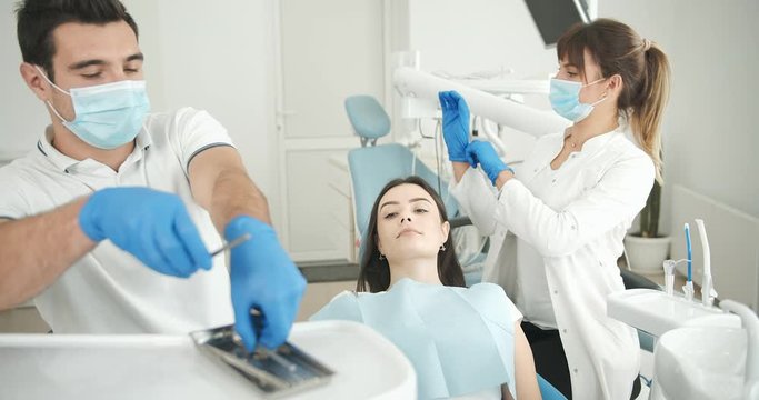 Young male dentist taking tools and checking woman's oral cavity, helpful assistant waiting near, stomatology