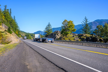 Big rig classic american bonnet semi truck with empty flat bed semi trailer driving on the road with rocks and autumn trees onthe sides