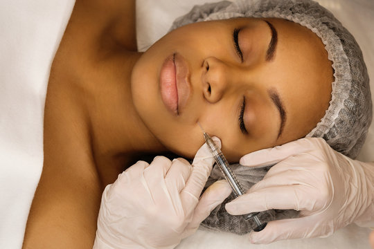 Injection cosmetology: contour plastic. Doctor makes rejuvenating facial injections procedure. Filling nasolabial fold with preparation with hyaluronic acid. Afro american woman receiving treatments