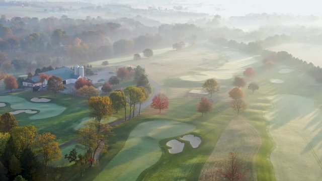 Cinematic high aerial reveals maintenance barn on golf course at sunrise, frost and fog in autumn sunlight