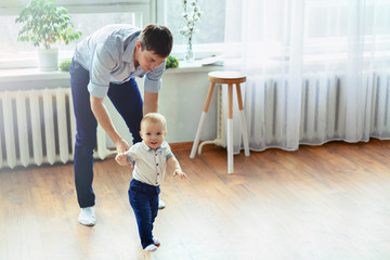 Daddy and son. Father in blue trousers and shirt teaches walk puny cheerful blond boy in bright...