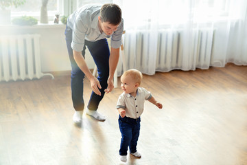 Daddy and son. Father in blue trousers and shirt teaches walk puny cheerful blond boy in bright...