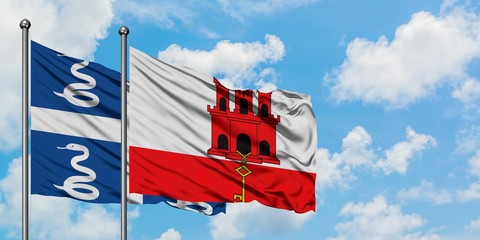Martinique and Gibraltar flag waving in the wind against white cloudy blue sky together. Diplomacy...