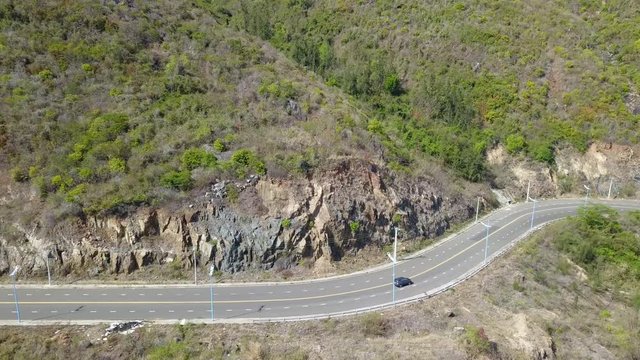 A flying camera takes a motorcyclist passing on a road near the sea with a view of the rocky shore.