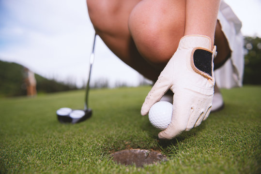 Female Golfers Pick Up Golf Balls In Holes On The Green Lawns. The Girl Friend Is Waiting For The Next Putt, Behind A Large Mountain. There Is Light From The Sunset.