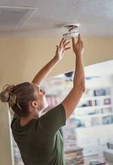 Changing the battery on a smoke alarm in an interior of a home. Woman standing on a step ladder and fixing the smoke alarm. © Brocreative
