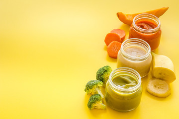 Baby food. Colorful puree in glass jars near vegetables and fruits on yellow background copy space