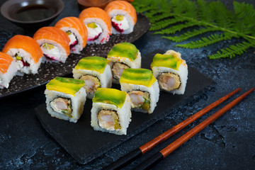 A set of sushi rolls with avocado and shrimp lies on a black charcoal Board. Traditional Japanese food with chopsticks