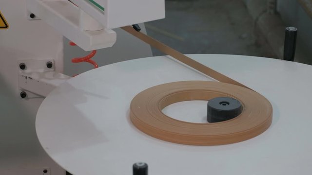 Furniture manufacture. Machine for edging chipboard parts. Pvc edge roll tension with wooden texture. Close-up. The video contains real production sound and noise.