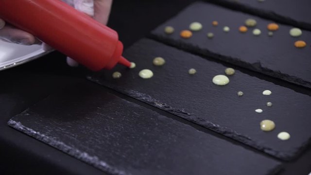 View of man's hands professionally decorating black rectangular plate by putting drops on it from yellow plastic bottle on table somewhere in restaurant preparing to catering.