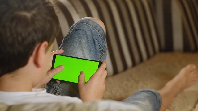 boy sits barefoot on couch at home in his room and holds smartphone with green screen and actively presses buttons with his fingers. child simulates the game in phone. Mobile phone with green screen.