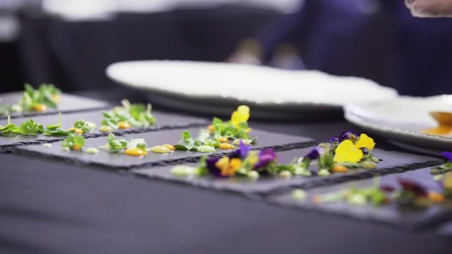 Camera shows man's hands making decoration of black rectangular plates by putting small colorful flowers on them on table with dark table cloth somewhere in restaurant preparing to catering.