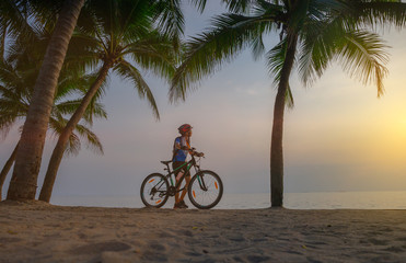 Fototapeta premium woman riding bicycle along the sea beach under coconut trees at sunset, relaxation at the end of the day