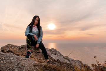 Naklejka premium The concept of Hiking and sports recreation. A woman with a backpack on her back sits on a rock and poses against the sea and sky. Sunset. Copy space