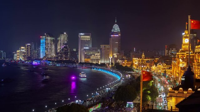 Timelapse famous Waitan waterfront area with illuminated Shanghai historical buildings at Huangpu river with Chinese flags on foreground at night