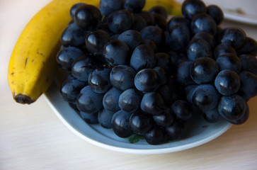 Bunches of fresh ripe red grapes and bananas on a white plate on wooden background. Tropical fruits. Healthy snack. Fruits, Healthy food concept, diet, healthy lifestyle.Ripe, fresh, juicy fruit.  