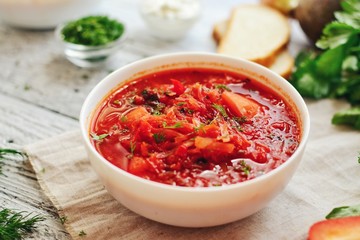 Appetizing Ukrainian and Russian borsch. Tomato soup in a white bowl. Vegan soup with tomato, sweet pepper, cabbage, carrots, beets and parsley. Food on a wooden background with textiles.