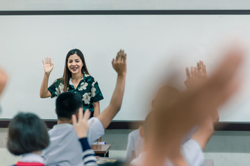 An smiling Asian female high school teacher teaches the white uniform students in the classroom by asking questions and then the students raise their hands for answers.