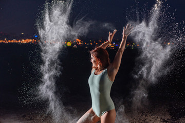 Red-haired woman in a blue bodysuit dancing in clouds of flour. A girl on the river bank jumping scattering white powder.