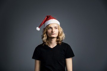 Young guy an attractive guy with long blonde hair in the form of a Christmas elf in a Santa Claus hat is standing with a displeased skeptical face on a gray background. Freelancer Party.