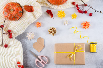 Gift box wrapped in craft paper, ribbon, bow, scissors and pumpkins and red berries on gray background. Thanksgiving, birthday presents preparation.