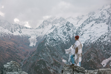 Fototapeta na wymiar Young guy wrapped in plaid blanket standing on mountain peak
