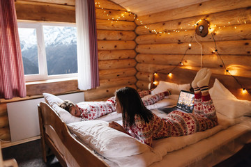 Teen friends in same Christmas pajamas relaxing in bed inside cozy log cabin with winter view