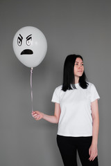 A girl in a white T-shirt with black hair holds in her hand a white balloon with an evil emotion.