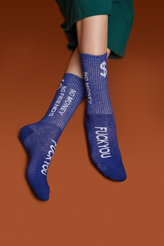 Close-up shot of female legs in wide green trousers and blue ribby socks with white lettering "No money. No friends. FuckYou". The photo is made on the brown background.