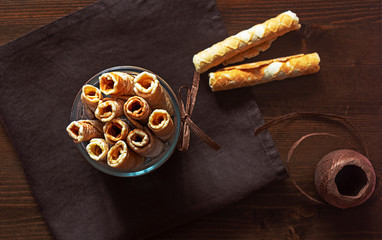 Tasty waffle rolls with icing sugar in a glass bowl with decorative decoration on a dark brown textile linen napkin on a wooden background. Sweet baked tubules and decorative ribbon for decoration. 