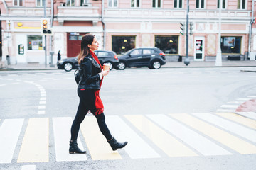 A beautiful brunette woman crosses the road on pedestrian crossing in a black leather jacket and a red scarf and hold in hand a hot drink.