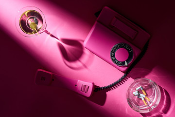 Top view of martini, pink telephone and astray with cigarette butts on pink surface