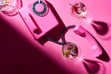Top view of cocktails and telephone handset beside astray with cigarette butts on pink surface