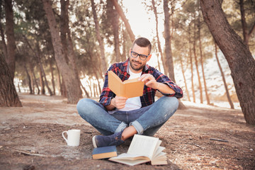 Hombre joven con lentes y camisa de cuadros, sentado en el suelo en medio del bosque, leyendo sonriente un libro al aire libre - obrazy, fototapety, plakaty