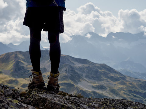 Silhouette of young hiker standing on cliff edge, legs view; brave man in with climbing equipment enjoying a freedom of ascent; mountain ranges and cloudy sky on background