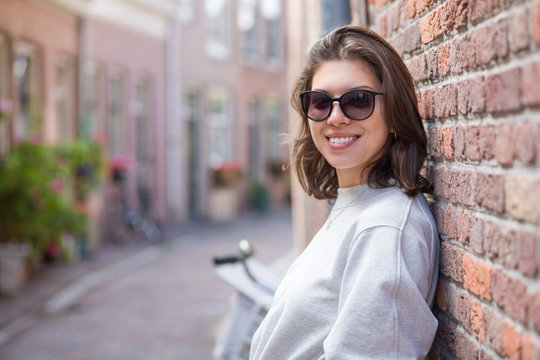 A happy young woman in sunglasses stands on a city street, leaning against a brick wall.Modern street fashion in the summer season.