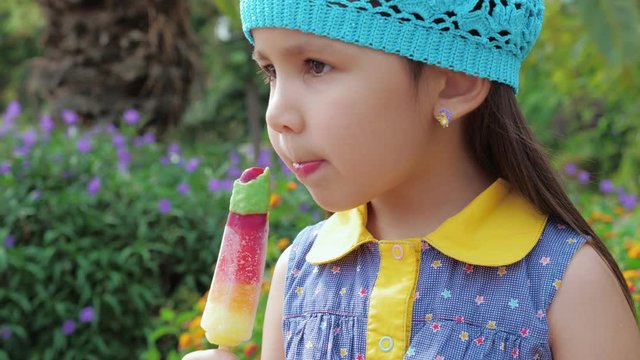 Cute little girl in a blue hat eating multicolored fruity ice cream. Trees and flowers at the background. Summer time.
