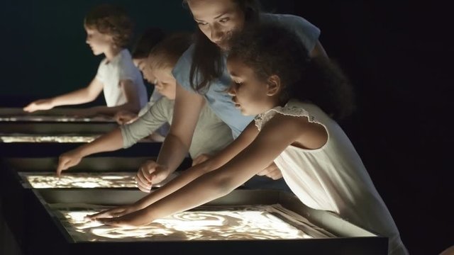 Tilt down shot of cheerful young female art teacher helping cute little girl making sand animation during group class in dark classroom