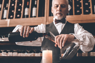 Sommelier Concept. Senior man standing pouring wine into decanter above candle concentrated bottom...