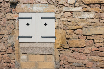 A fragment of an old wall made of rough stones of various shapes and sizes. There is a window closed by white shutters. Europe. France. Background. Texture.
