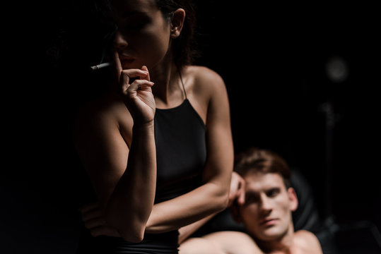 seductive young couple sitting on sofa in dark room, while sexy woman smoking cigarette