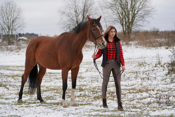 A beautiful girl in a fashionable shirt walks with broun horse in early winter.