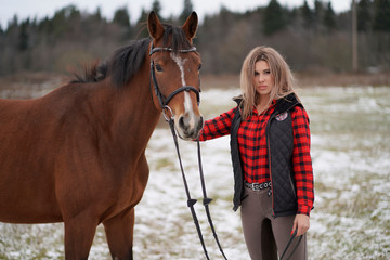 A beautiful girl in the red plaid shirt walks with broun horse in early winter.